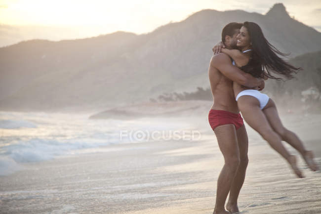 Casal adulto médio na praia, vestindo roupa de banho, homem balançando mulher rodada — Fotografia de Stock