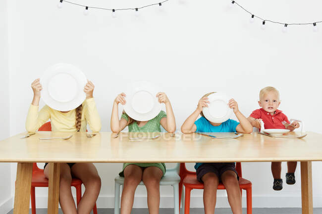 Cuatro niños sentados a la mesa, tres caras cubiertas con platos - foto de stock