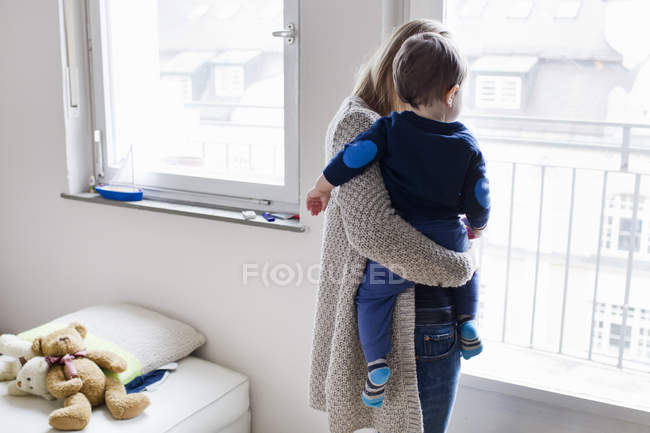 Vue arrière de la femme adulte moyenne et du bébé fils regardant par la fenêtre — Photo de stock