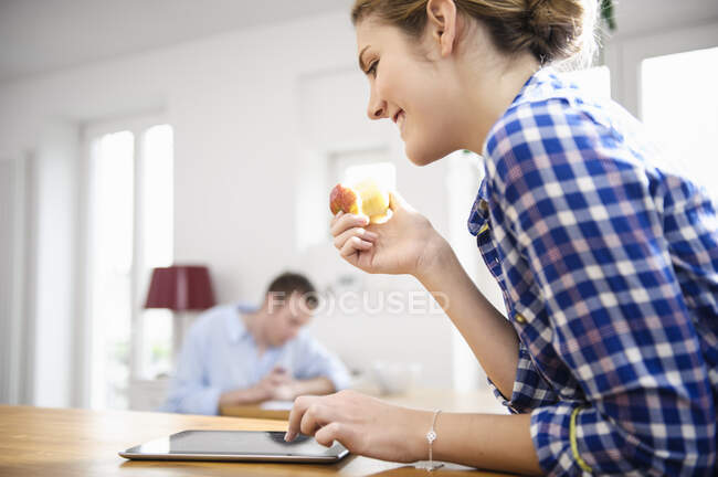 Junge Frau im blau karierten Hemd isst einen Apfel und benutzt ein Tablet — Stockfoto