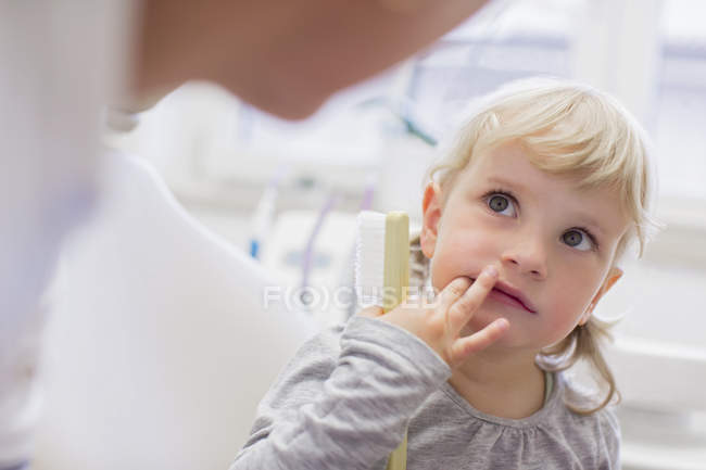 Chica con dedos en la boca sosteniendo el cepillo de dientes mirando al dentista - foto de stock