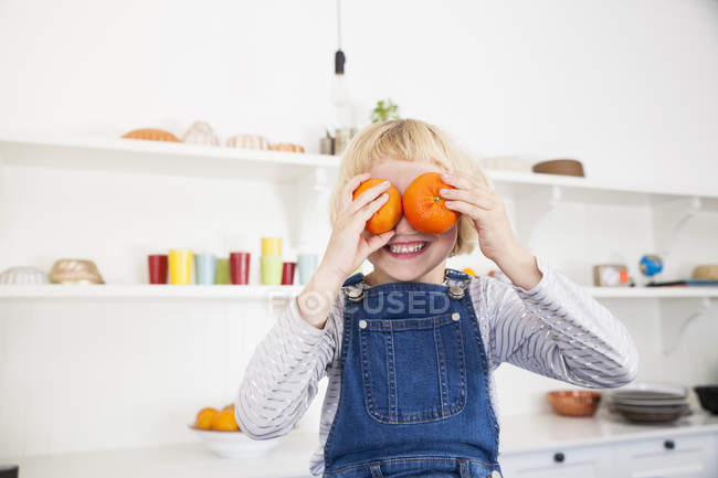 Portrait of cute girl in kitchen holding oranges to her eyes — Stock Photo