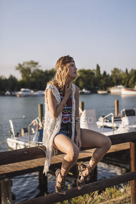Blonde Caucasian woman sitting on fence near lake water with boats and yachts — Stock Photo
