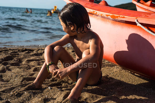 Мальчик сидит на пляже на лодке и смотрит в сторону — стоковое фото