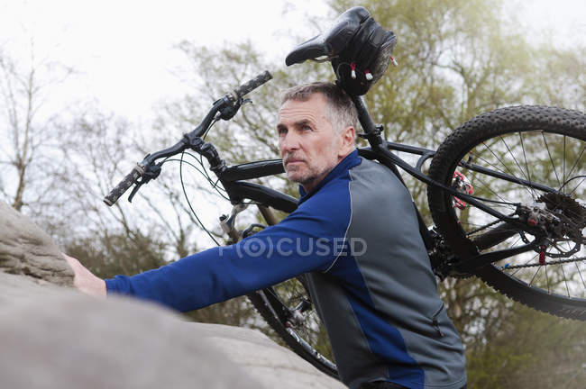 Mature male mountain biker carrying bike over rock formation — Stock Photo