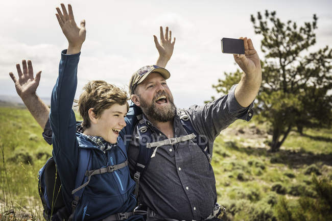 Padre e hijo adolescente saludando para selfie smartphone en viaje de senderismo, Cody, Wyoming, EE.UU. - foto de stock