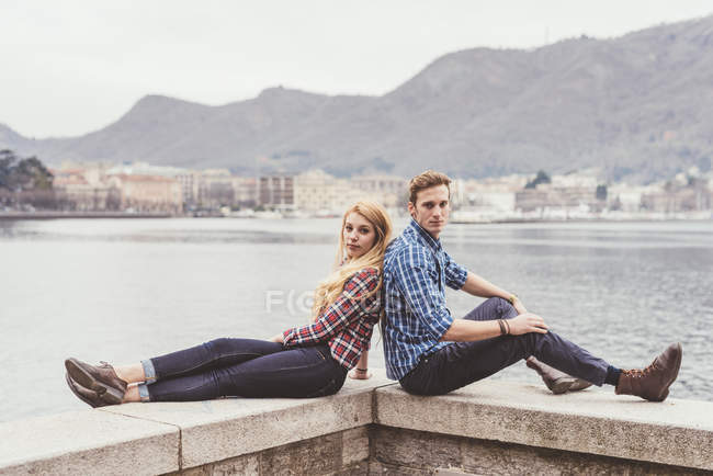 Портрет молодой пары, сидящей спиной к спине на стене гавани, озеро Комо, Италия — стоковое фото
