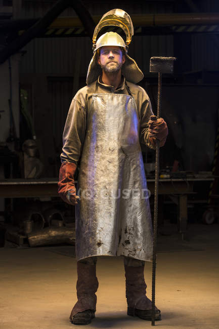 Retrato del trabajador de fundición macho maduro sosteniendo rastrillo en fundición de bronce - foto de stock