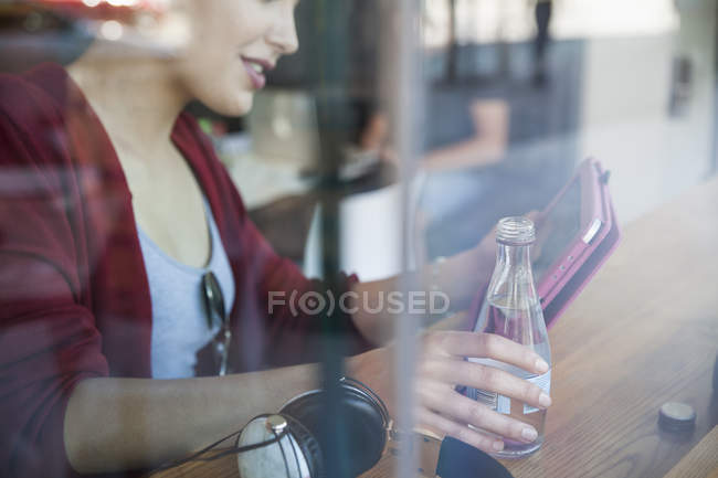 Молода жінка сидить у кафе, тримає пляшку води, використовуючи цифровий планшет — стокове фото