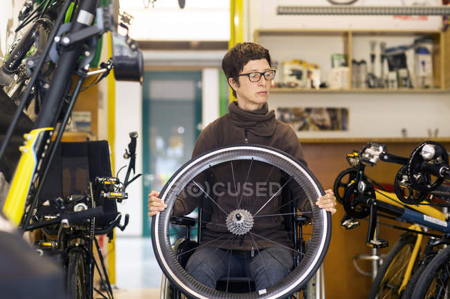 Woman in wheelchair in bicycle repair shop, holding bicycle wheel — Stock Photo