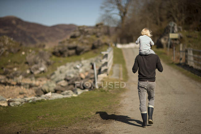 Padre llevando a su hijo en hombros, caminando por el camino, vista trasera - foto de stock