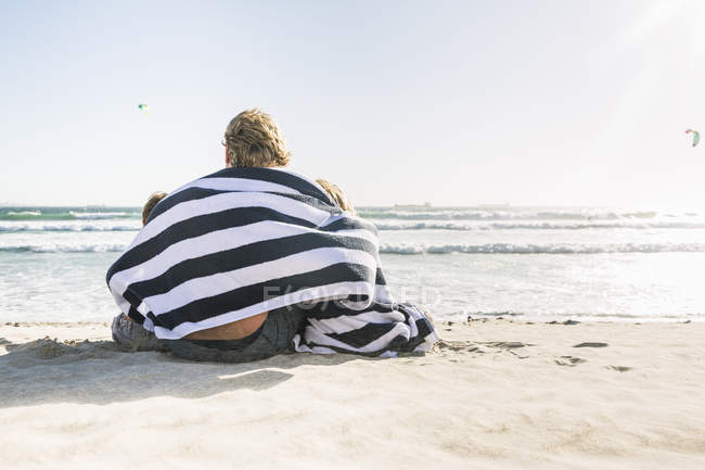Vista posteriore di padre e figli seduti sulla spiaggia avvolti in una coperta — Foto stock
