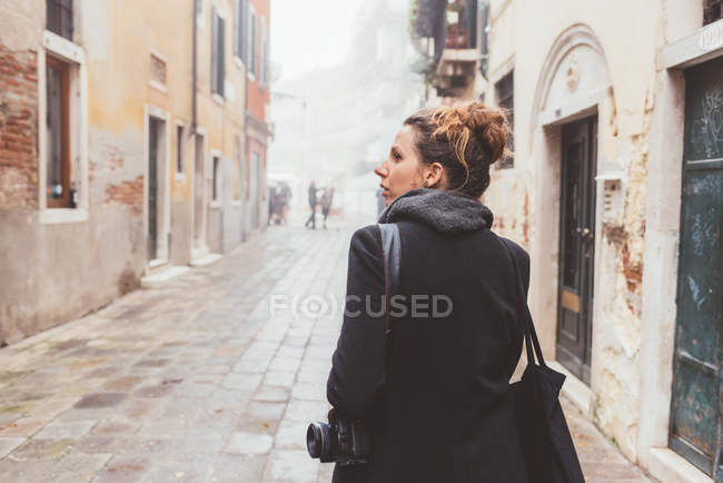 Mujer joven con cámara mirando por encima de su hombro en la calle, Venecia, Italia - foto de stock
