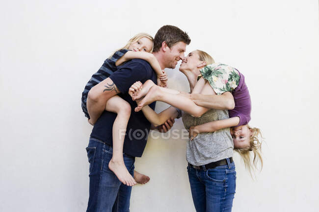 Portrait of parents carrying two young daughters in front of white wall — Stock Photo