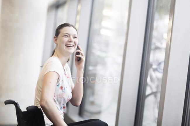 Молодая женщина в инвалидной коляске у входной двери разговаривает на смартфоне — стоковое фото