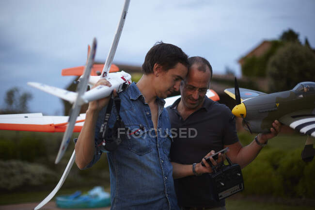 Deux amis masculins tenant des avions télécommandés, regardant smartphone — Photo de stock