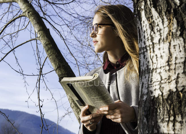 Mujer joven de pie junto al árbol, sosteniendo el libro - foto de stock