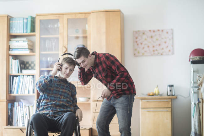 Молодой человек на инвалидной коляске слушает музыку в наушниках с другом на кухне — стоковое фото