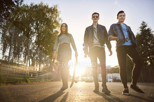 Portrait of young female skateboarder and male friends walking on sunlit street — Stock Photo