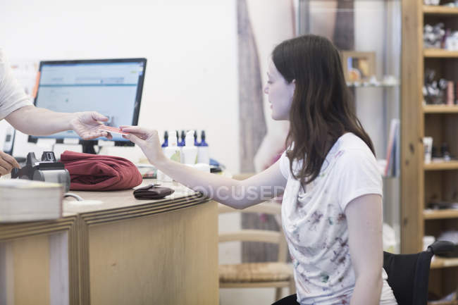 Jeune femme utilisant un fauteuil roulant payer avec une carte de crédit dans la boutique — Photo de stock