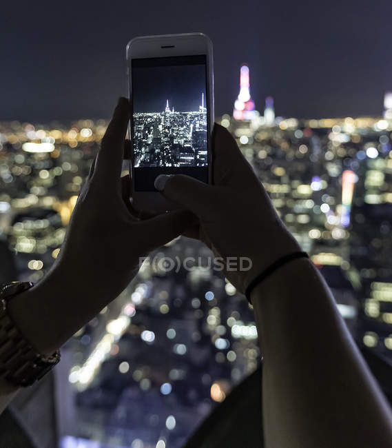 Manos femeninas con teléfono inteligente tomar fotos de la noche iluminado paisaje urbano de Nueva York - foto de stock