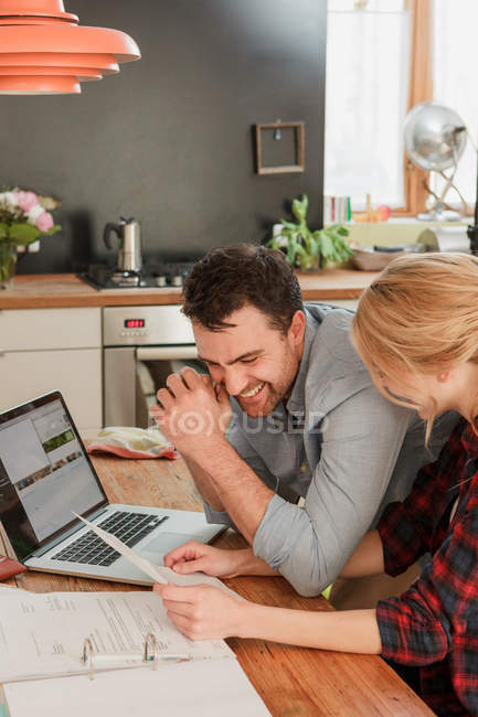Casal na mesa de jantar com laptop olhando papelada sorrindo — Fotografia de Stock