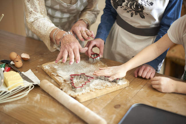 Foto recortada de mujer mayor y nietas cortando galletas de árbol de Navidad en el mostrador de la cocina - foto de stock