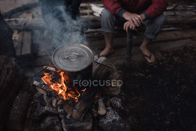 Homem sênior sentado no log, assistindo panela de cozinha na fogueira, seção baixa — Fotografia de Stock
