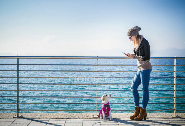 Полный вид сбоку молодой женщины с собакой за перила перед океаном с помощью смартфона — стоковое фото