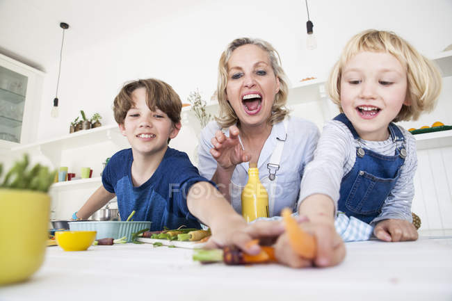 Niña y hermano buscando vegetales mientras preparan comida en la mesa de la cocina con la madre - foto de stock