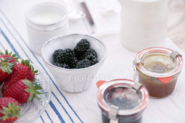 Summer fruits in bowls and preserves in jars on tablecloth — Stock Photo