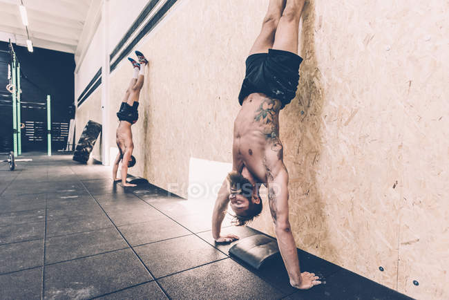 Two young male cross trainers doing handstands against gym wall — Stock Photo