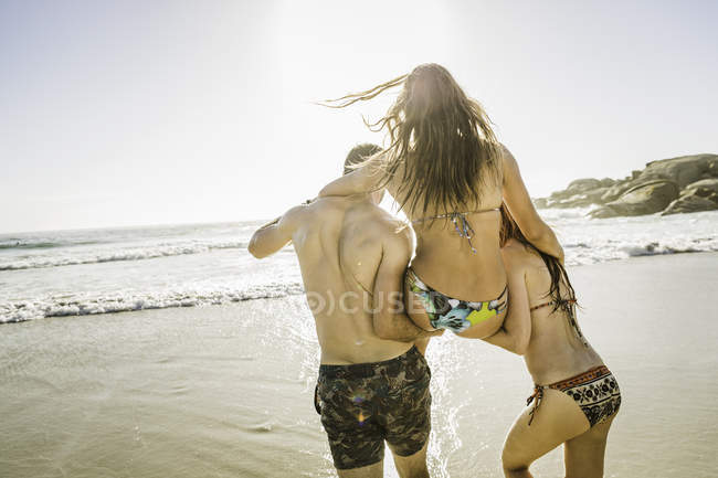 Вид сзади на пару, несущую женщину в бикини на пляже, Кейптаун, ЮАР — стоковое фото