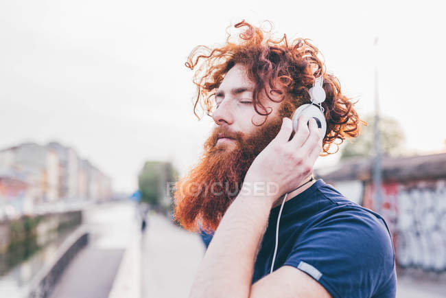 Young male hipster with red hair and beard listening to headphones with eyes closed in city — Stock Photo