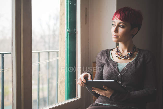 Молода жінка сидить біля вікна, використовуючи цифровий планшет — стокове фото