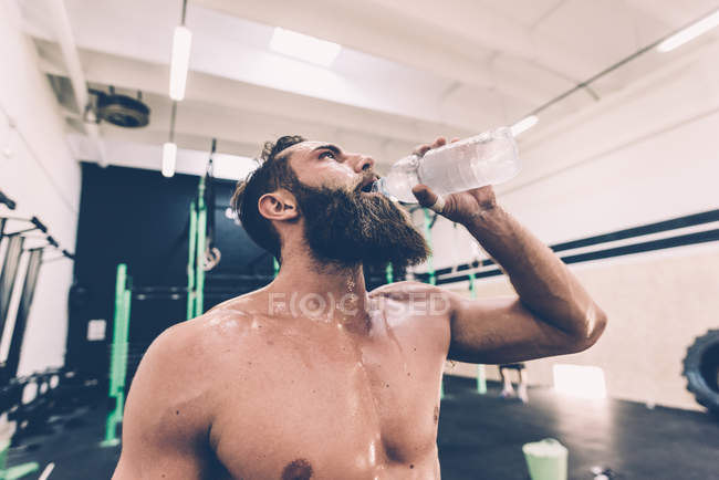 Entrenador cruzado masculino agotado bebiendo agua en el gimnasio - foto de stock