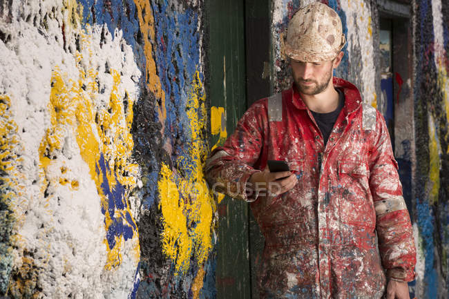 Männlicher Schiffsmaler liest Smartphone-Text, der sich gegen eine mit Farbe bespritzte Wand lehnt — Stockfoto