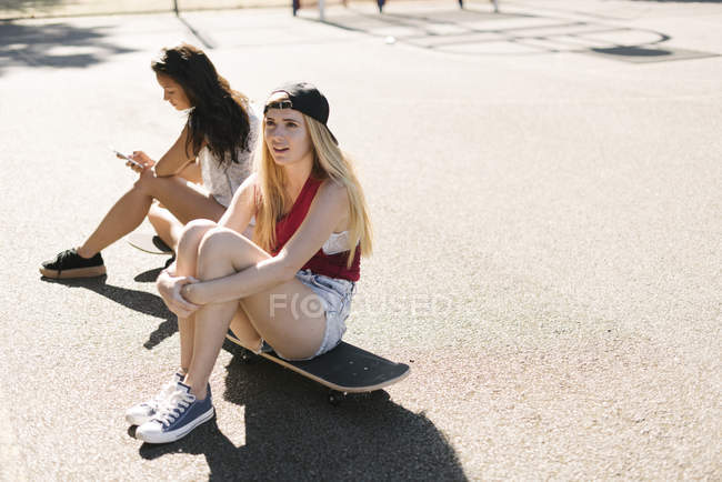 Two adult female friends sitting on skateboards on basketball court — Stock Photo