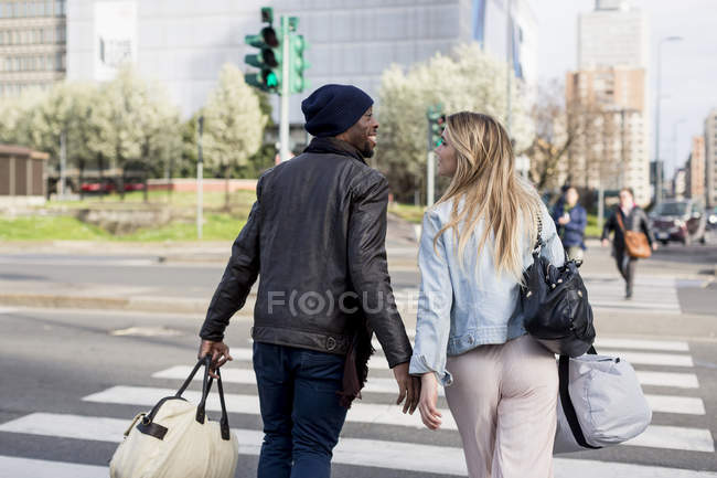 Щаслива усміхнена багатонаціональна пара перетинає дорогу на вулиці і переносить сумки — стокове фото