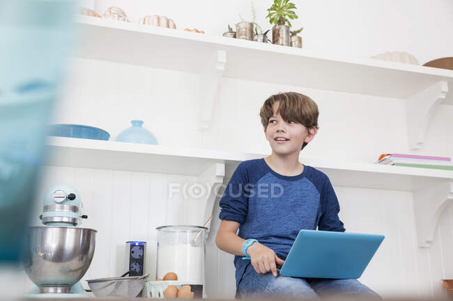 Menino sentado na superfície de trabalho da cozinha, usando computador portátil — Fotografia de Stock