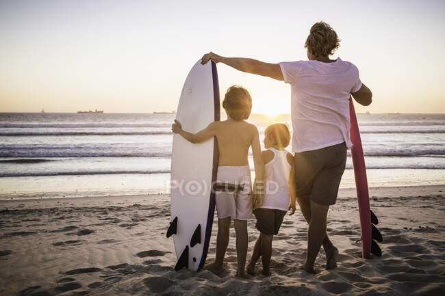 Padre y dos hijos de pie en la playa, con tablas de surf, mirando al océano, vista trasera - foto de stock