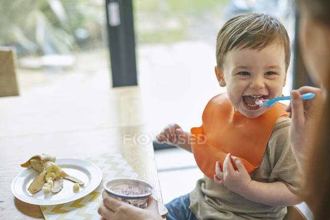 Happy baby boy sitting at table being fed yogurt by mother — Stock Photo