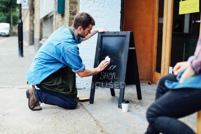 Young man kneeling on pavement and wiping blackboard outside workshop — Stock Photo