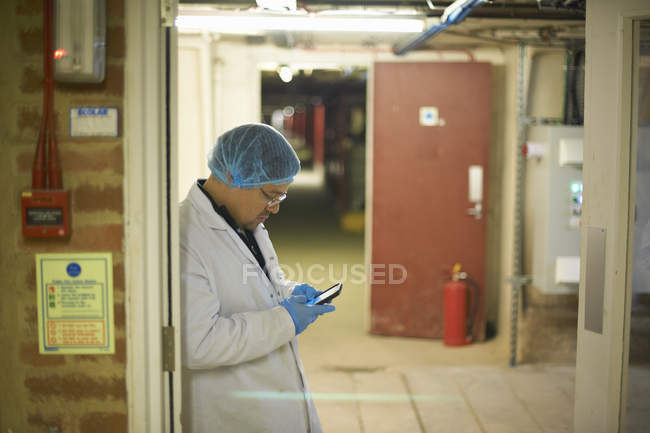 Worker wearing hair and latex gloves leaning against doorway using smartphone — Stock Photo
