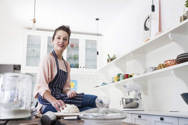 Portrait of young woman sitting on kitchen counter preparing dough — Stock Photo