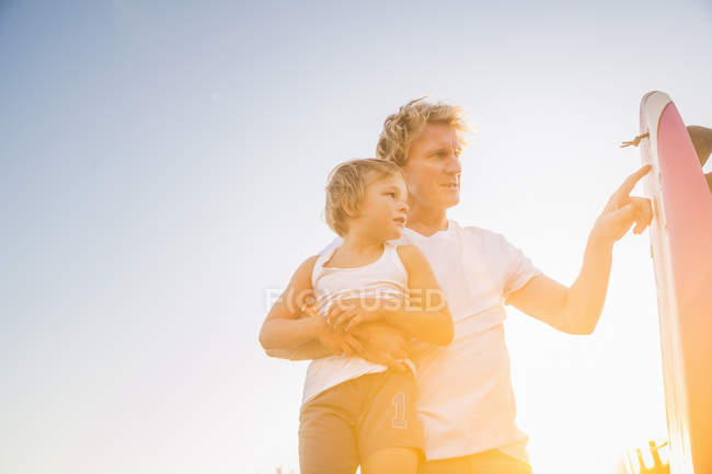 Vista de ángulo bajo del padre sosteniendo al hijo mirando la tabla de surf - foto de stock