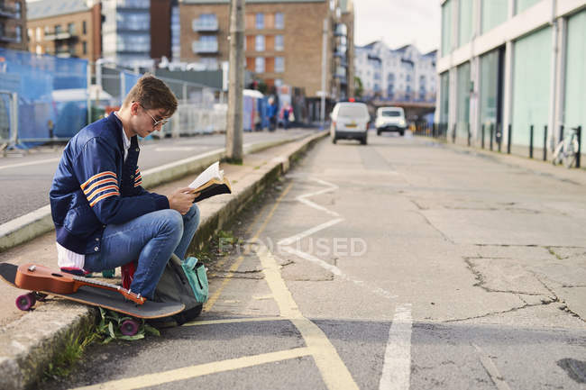 Young man sitting on kerb, reading book, skateboard beside him, Bristol, UK — Stock Photo