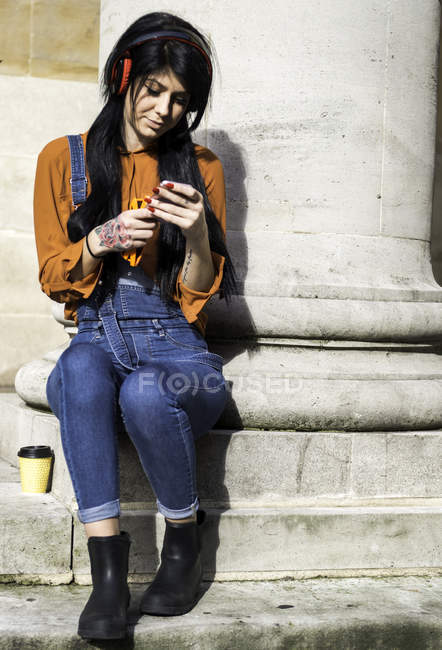 Молодая женщина сидит у столба, в наушниках, держит смартфон — стоковое фото