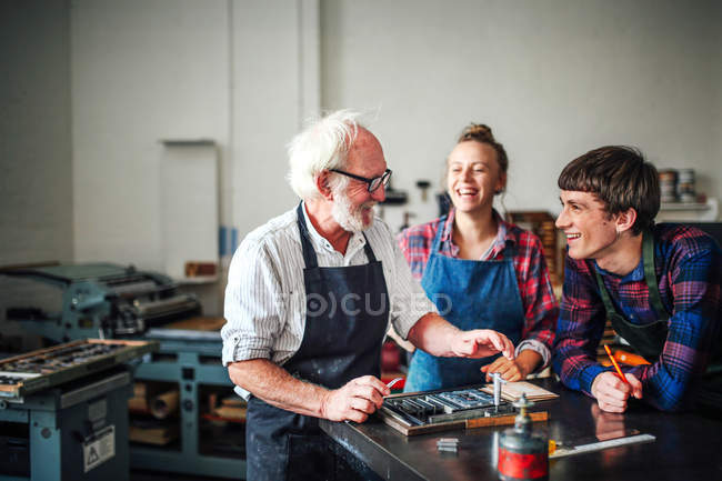 Artesano senior riéndose con joven artesano y artesana en taller de tipografía - foto de stock
