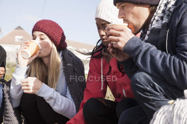 Amigos adultos jóvenes agachados en la playa tomando café para llevar, Western Cape, Sudáfrica - foto de stock